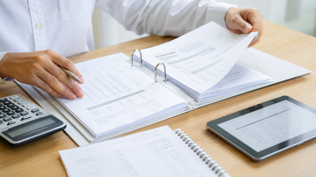 Business professional reviewing financial documents and reports in a binder during strata consultation, with calculator and tablet on wooden desk