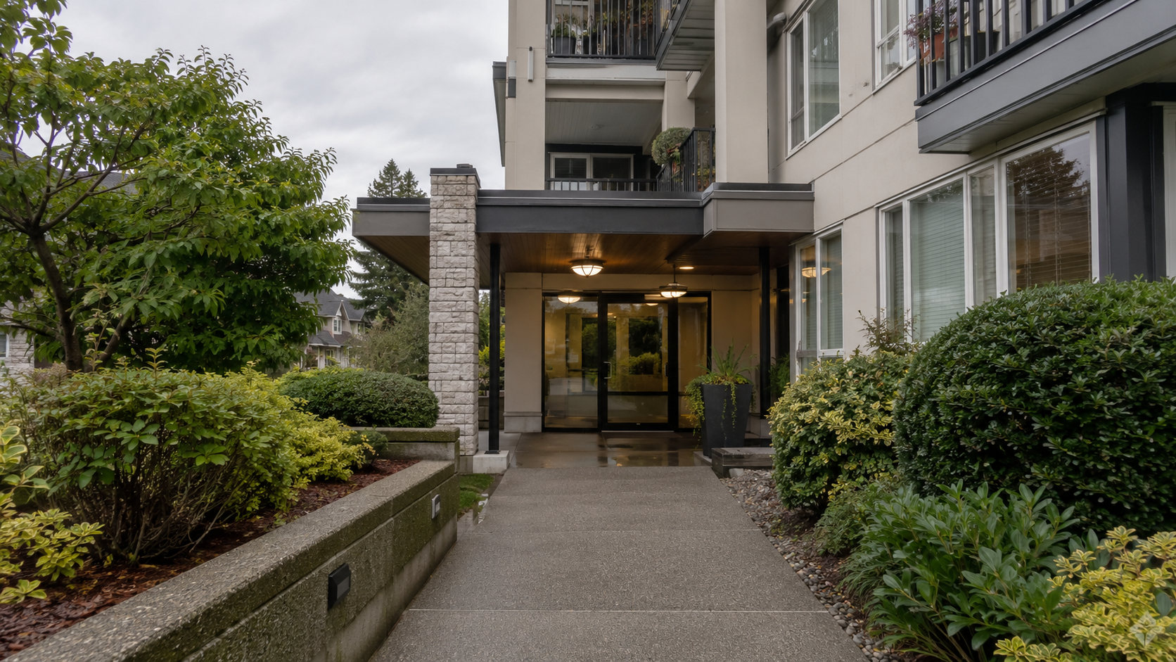 Modern residential building entrance with paved pathway, professional landscaping with hedges and shrubs, and glass doors leading to the lobby