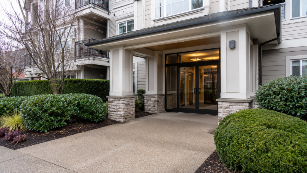 Modern apartment building entrance with clean concrete walkway, manicured hedges, and glass entry doors - representing professional strata property management services for self-managed strata corporations in Surrey BC