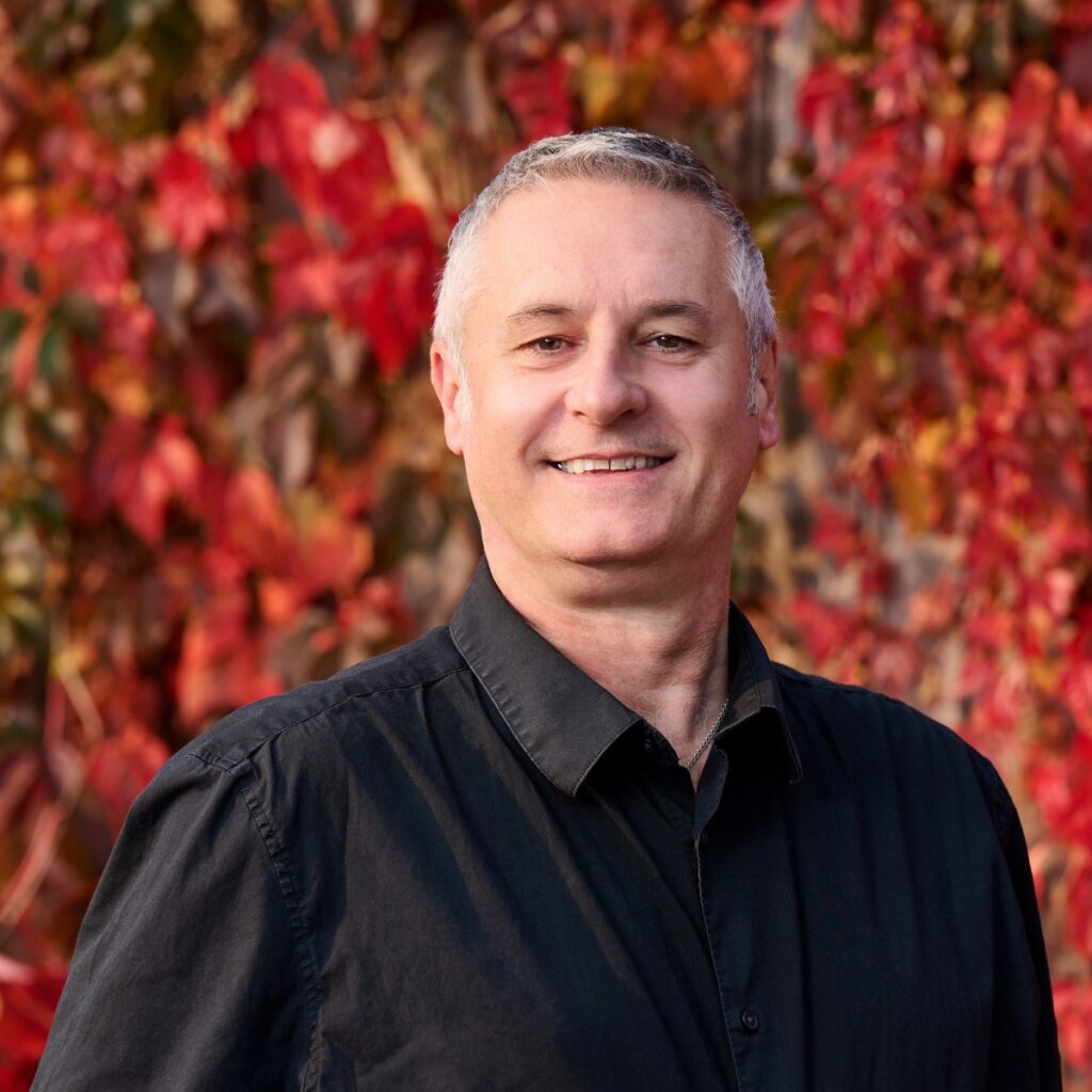 Portrait of smiling man in black shirt standing in front of red autumn leaves, representing All Property Consulting's team for self-managed strata in Surrey BC