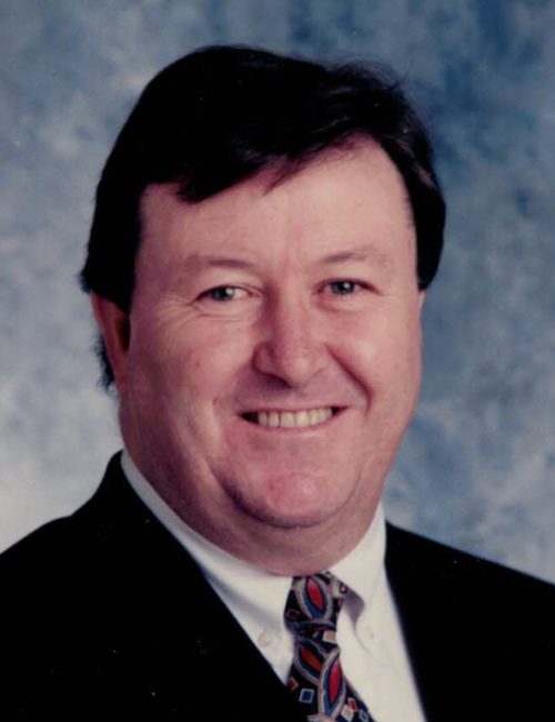 Professional headshot of a smiling man in a dark suit and patterned tie against a blue background